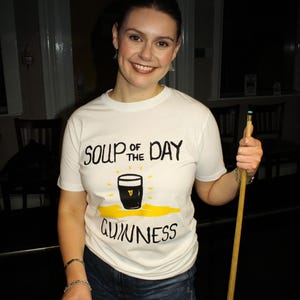 以下が含まれることがあります： 「Soup of the Day Guinness」の文字とギネスビールのパイントのグラフィックがプリントされた白いTシャツ。ビリヤードのキューとギネスビールのパイントを持っています。青いジーンズを着用しています。