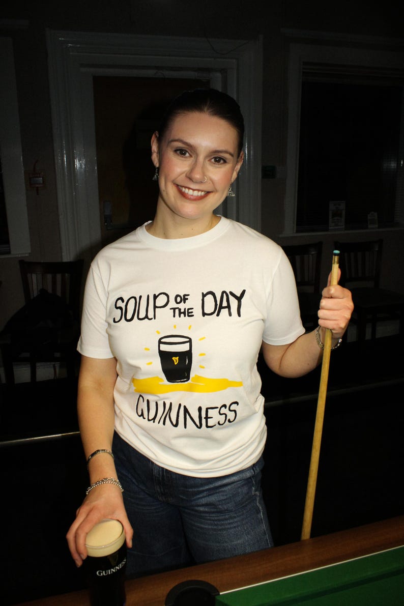 以下が含まれることがあります： 「Soup of the Day Guinness」の文字とギネスビールのパイントのグラフィックがプリントされた白いTシャツ。ビリヤードのキューとギネスビールのパイントを持っています。青いジーンズを着用しています。