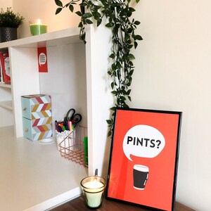 Puede incluir: Un cartel enmarcado con la palabra "PINTS?" en un bocadillo sobre una pinta de cerveza negra sobre un fondo rojo. El cartel se muestra sobre una superficie de madera, junto a una vela encendida y una planta decorativa.