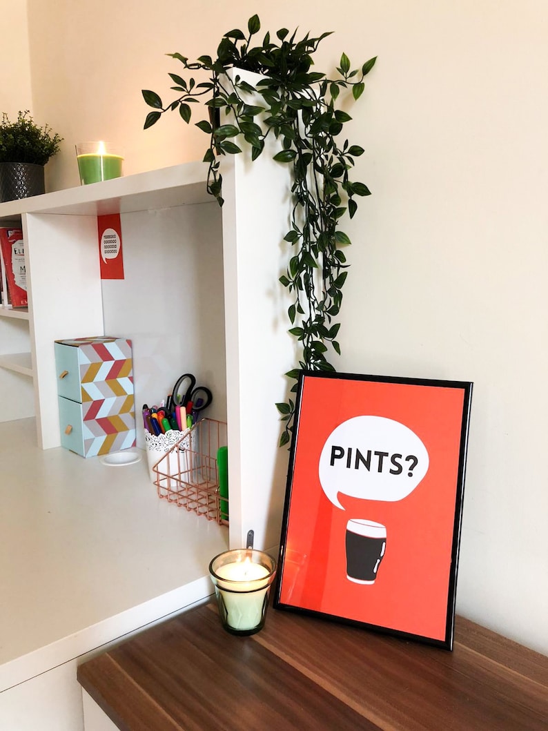 Puede incluir: Un cartel enmarcado con la palabra "PINTS?" en un bocadillo sobre una pinta de cerveza negra sobre un fondo rojo. El cartel se muestra sobre una superficie de madera, junto a una vela encendida y una planta decorativa.