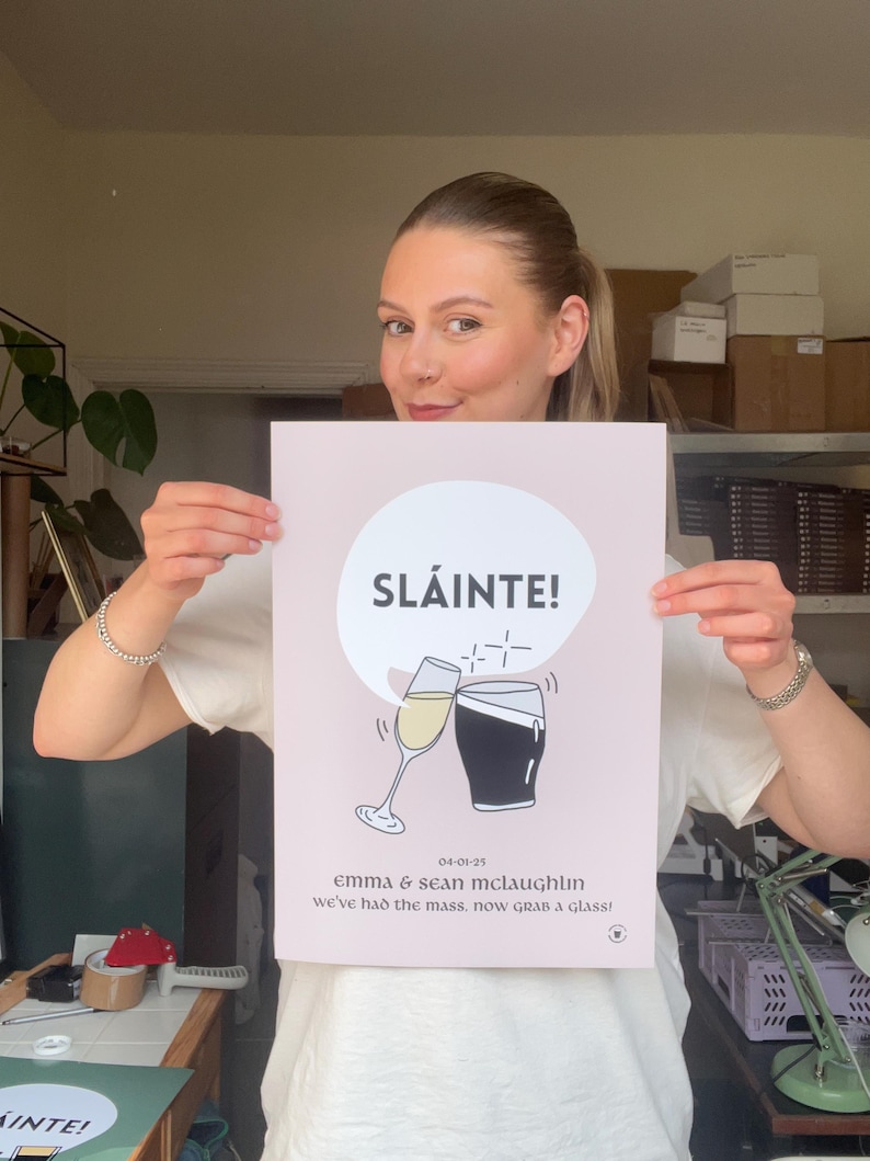 Op de afbeelding: Een roze poster met het woord "SL&Aacute;INTE!" in een witte cirkel, met illustraties van een champagneglas en een pint stout die klinken. De poster bevat de datum 04-01-25 en de namen Emma & Sean McLaughlin.