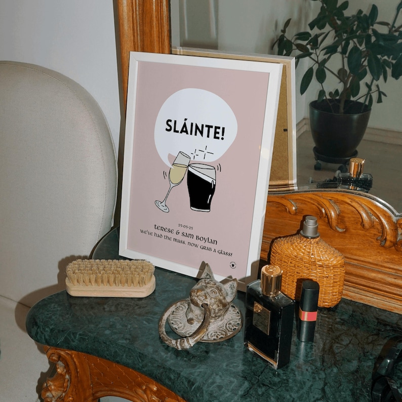 Op de afbeelding: Een ingelijste print met het woord "SL&Aacute;INTE!" en een afbeelding van twee klinkende glazen. Onder de afbeelding staat de tekst "Terese & Sam Boylan" en "we've had the mass, now grab a glass!". De print wordt tentoongesteld op een marmeren oppervlak.