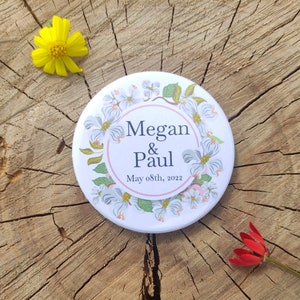 May include: White round button with a floral design and the text "Megan & Paul May 08th, 2022" in black.
