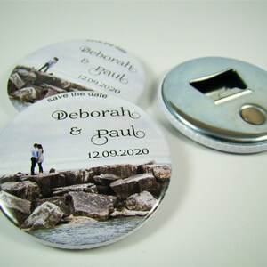 May include: Two round, white, magnetic bottle openers with a photo of a couple on a rocky beach. The text "Deborah & Paul 12.09.2020 save the date" is printed on the opener.