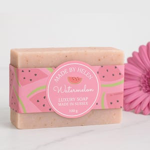 May include: A rectangular bar of luxury soap with a pink and green watermelon-themed label. The label reads "Made by Helen Watermelon Luxury Soap Made in Sussex 100 g". A pink flower is in the background.