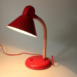 Könnte beinhalten: Eine rote Schreibtischlampe mit flexiblem Hals und einer weißen Glühbirne. Die Lampe hat einen runden Sockel und einen nach unten geneigten Schirm.