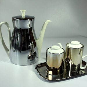 May include: A silver coffee pot with a white lid and a silver tray with two silver creamer pitchers with white lids.