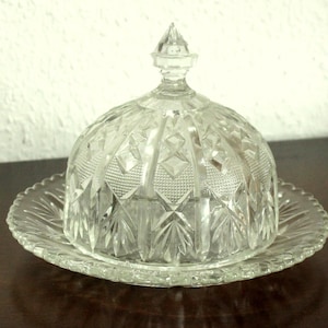 Op de afbeelding: Een heldere, vintage glazen botervloot met een gewelfde deksel en een decoratieve, geschulpte randplaat. De deksel heeft een puntige finial en een ruitpatroon. De schaal is ontworpen om boter in te bewaren en is een klassiek keukenartikel.