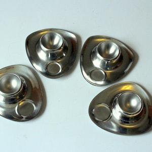 May include: Four stainless steel egg cups with a modern, triangular design. Each cup has a central holder for the egg and a small, circular indentation. The reflective surface of the cups creates highlights.