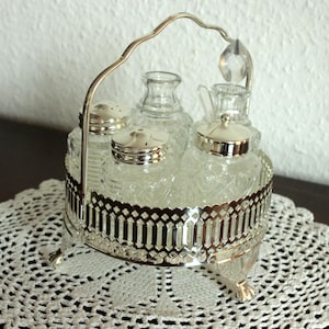 Op de afbeelding: Een vintage zilverkleurige kruidenset met heldere glazen flessen en een decoratieve metalen houder. De set bevat twee kleine shakers en twee flessen met deksels, allemaal rustend op een decoratieve, opengewerkte basis.