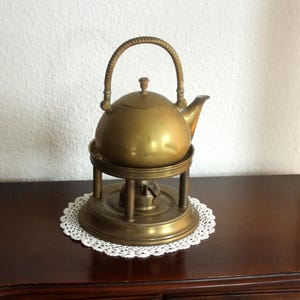 Op de afbeelding: Een vintage messing theepot met een gevlochten handvat en een kleine knop op het deksel. De theepot staat op een bijpassende messing standaard met een brander, rustend op een wit kanten kleedje. De set wordt gepresenteerd op een donker houten oppervlak.