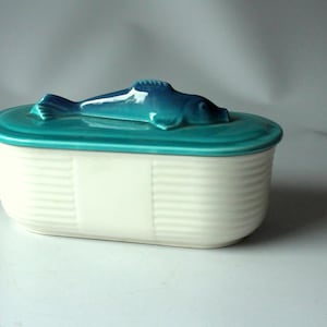 Może przedstawiać: Ceramiczny pojemnik z pokrywką z turkusową rzeźbą ryby. Pokrywka i ryba są w żywym turkusie, kontrastując z białym, żebrowanym korpusem pojemnika. Z przodu znajduje się prostokątna biała przestrzeń.