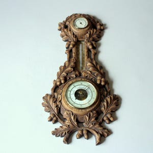 May include: A wooden weather station with a barometer, thermometer, and hygrometer. The decorative piece is carved with oak leaves and acorns. The barometer dial has numbers and a needle. The thermometer is a glass tube with a scale. The hygrometer is a small dial.