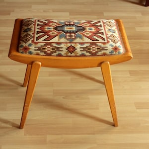 Peut inclure: Un pouf en bois vintage avec une assise rectangulaire. L'assise est recouverte d'un tissu à motifs colorés dans les tons de rouge, bleu et beige. Le pouf a quatre pieds inclinés et un cadre incurvé.