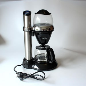 Könnte beinhalten: Eine schwarze Elta Kaffeemaschine aus Edelstahl mit einem hohen zylindrischen Turm, einem klaren Glaswasserbehälter mit schwarzem Deckel, einem zentralen Filterkorb und einer Glaskaraffe mit schwarzem Griff, verbunden mit einem schwarzen Stromkabel.
