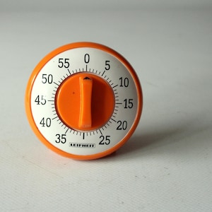 Puede incluir: Un temporizador de cocina redondo de color naranja y blanco con un dial central naranja. La esfera blanca muestra números negros del 0 al 55 en incrementos de cinco, con marcas para los minutos. La marca LEIFHEIT está impresa en negro abajo.