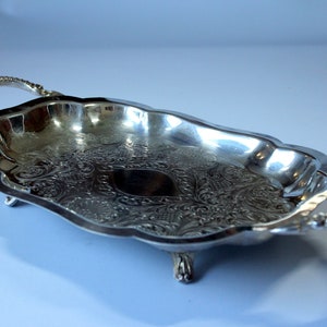 Könnte beinhalten: Ein silbernes Serviertablett mit verzierten Details und zwei Griffen. Das Tablett hat drei Füße und ist oval.