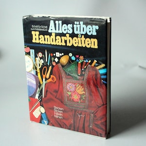 May include: A vintage book titled "Alles über Handarbeiten" (All About Handicrafts) with a black cover and colorful illustrations. The cover features text and images related to sewing, knitting, and other crafts. The book is in German.