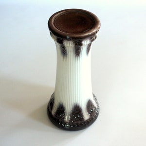 Op de afbeelding: Een decoratieve vaas in wit en bruin. De cilindrische vaas heeft verticale geribbelde details. De boven- en onderkant zijn bruin, met een gestructureerd, bloemachtig patroon aan de basis. Een stijlvol woonaccessoire.