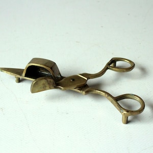 Könnte beinhalten: Ein Dochtschneider aus Messing mit scherenartigem Design, ausgestattet mit zwei Fingerringen, einem zentralen Drehpunkt und einem rechteckigen, kastenförmigen Aufsatz an einer Klinge, der dazu dient, abgeschnittene Dochtstücke aufzufangen.