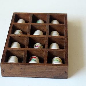 Könnte beinhalten: Eine Holzvitrine mit zwölf Fächern, in denen jeweils ein weißer Fingerhut aufbewahrt wird. Mehrere Fingerhüte weisen florale Designs in verschiedenen Farben auf. Die Vitrine ist aus dunkelbraunem Holz gefertigt.