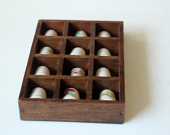 May include: A wooden display case with twelve compartments, each holding a white thimble. Several thimbles feature floral designs in various colors. The case is made of dark brown wood.
