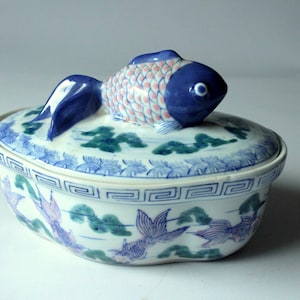 Könnte beinhalten: Ovale Keramikschale mit Deckel, mit einem blauen Fischgriff. Die Schale ist weiß mit blauen und grünen Blumenmustern und einem geometrischen Rand. Der Fisch hat blaue und rosa Schuppen.