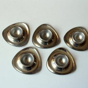 May include: Five silver-colored egg cups with a unique, triangular design. Each cup has a rounded interior to hold an egg. The egg cups are made of a shiny metal and are arranged on a white surface.