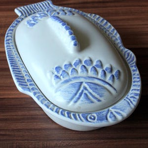 May include: A white ceramic lidded dish shaped like a fish, featuring embossed scale patterns and a raised dorsal fin handle. The edges and decorative details are accented with blue paint, creating a textured appearance on the lid and rim.