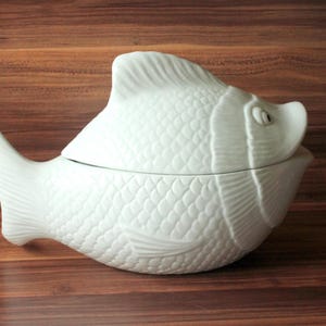 Może przedstawiać: Białe ceramiczne naczynie do serwowania w kształcie ryby z pokrywką. Naczynie ma szczegółowy wzór łusek i detale płetw. Naczynie stoi na drewnianej powierzchni.