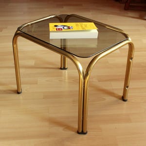 Könnte beinhalten: Ein rechteckiger Vintage-Couchtisch mit einer Rauchglasplatte und einem goldfarbenen Metallrahmen. Ein Buch mit gelbem Einband liegt auf der Glasoberfläche. Der Tisch ist ca. 50 cm lang, 38 cm breit und 46 cm hoch.