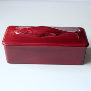 Könnte beinhalten: Eine rechteckige, rubinrote Keramikbox mit Deckel. Der Deckel zeigt ein erhabenes Relief eines Fisches. Die Box hat eine einfarbige, glänzende Farbe und steht auf einer weißen Oberfläche. Die Box dient wahrscheinlich zur Aufbewahrung oder Dekoration.