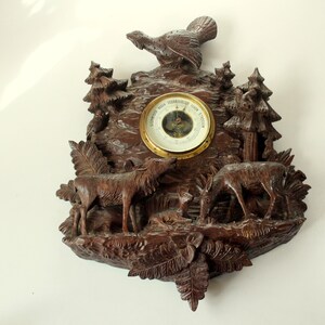 May include: A dark brown, carved wooden wall barometer featuring a forest scene. The scene includes deer, trees, and a bird. The barometer has a gold-colored bezel and a white face with black markings.