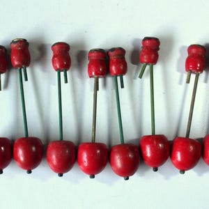 May include: A collection of eight wooden cherry ornaments. Each ornament features a bright red cherry with a small black dot, a green or brown stem, and a smaller red top. The ornaments are arranged in a row.