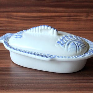 Może przedstawiać: Białe ceramiczne naczynie do serwowania z pokrywką, z niebieskimi wzorami kwiatowymi i geometrycznymi. Naczynie ma uchwyt na pokrywce i ozdobną obwódkę. Tło to ciemna drewniana powierzchnia.