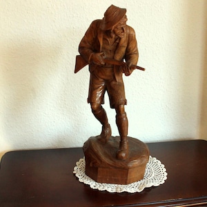 Könnte beinhalten: Eine Holzskulptur eines Jägers, der ein Gewehr hält und auf einem Holzsockel steht. Die Skulptur ist detailliert geschnitzt und wirkt realistisch.