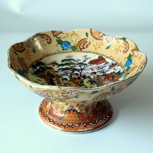 May include: A decorative ceramic bowl with a pedestal base. The bowl is decorated with a colourful scene of people in a Japanese garden. The bowl is cream-coloured with gold accents.