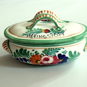 Könnte beinhalten: Ovale Servierschale aus Keramik mit Deckel. Die Schale ist weiß mit grünen Verzierungen und einem Griff. Sie ist mit bunten Blumen- und Blattmotiven verziert. Das Wort "HERINGS STOPF" steht auf der Seite.
