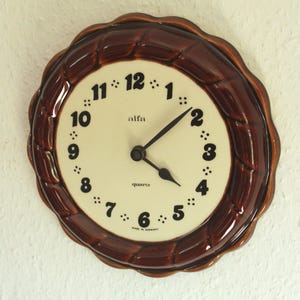 Könnte beinhalten: Runde Wanduhr mit dunkelbraunem Rahmen und cremefarbenem Zifferblatt. Schwarze Ziffern und Zeiger. Über der Mitte steht "alfa", darunter "quartz". Aufschrift "Made in Germany".