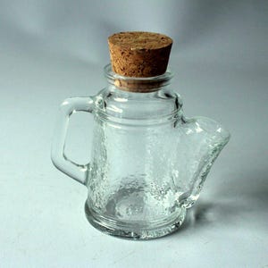 Peut inclure: Une bouteille en verre transparent en forme de théière, avec une surface texturée et un bouchon en liège. La bouteille a une poignée et un bec verseur, ressemblant à une théière miniature. Le bouchon est brun clair.