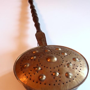 May include: A vintage copper and wood stove top kettle with a long wooden handle and a perforated bottom. The kettle has a decorative design of raised circles and dots.