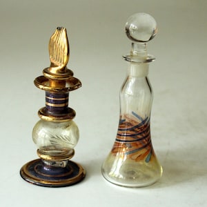 Könnte beinhalten: Zwei dekorative Glasparfümflakons mit goldenen Akzenten. Die Flasche links hat einen goldenen und violetten Boden und einen goldenen Deckel mit einem spitzen Abschluss. Die Flasche rechts hat einen durchsichtigen Glaskugelverschluss und einen durchsichtigen Glaskörper mit goldenen und blauen Wirbeln.