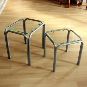 May include: Two modern nesting tables with clear glass tops and silver metal frames. The tables are different sizes, with the larger one on the left and the smaller one on the right. The tables are on a light wood floor.