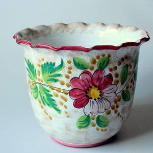 Könnte beinhalten: Keramik-Blumentopf mit gewelltem Rand. Der Topf ist mit einem Blumendesign bemalt, das eine große rosa Blume, grüne Blätter und gelbe Punkte zeigt. Der Rand und der Boden sind rot bzw. rosa eingefasst.