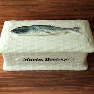 Peut inclure: Une boîte en céramique blanche avec un ornement en forme de poisson sur le couvercle. La boîte présente un motif de panier tressé et le texte "Marin. Heringe" sur le devant. Le poisson est gris et blanc.