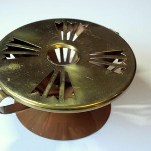 Könnte beinhalten: Ein Vintage-Kerzenwärmer aus Messing und Kupfer. Die runde Oberseite hat dekorative Aussparungen und einen Griff. Der Sockel hat eine konische Form aus Kupfer. Der Kerzenwärmer steht auf einer weißen Oberfläche.