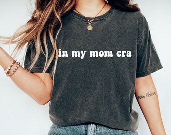 May include: A dark gray t-shirt with the text "in my mom era" printed in white. The text is in a retro font style.