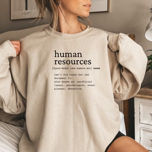 May include: A beige sweatshirt with the text "human resources" and a definition of the term. The definition includes the pronunciation, "[hyoo-muhn ree-sawrs-es]", and describes the role as "can't fix crazy but can document it." It also lists "unofficial lawyer, psychologist, event planner, detective" as other names for the role.