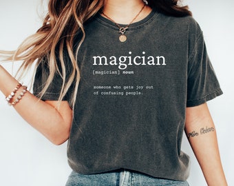 May include: A dark gray t-shirt with the word "magician" printed in white. The definition of the word is printed below: "[magician] noun someone who gets joy out of confusing people."