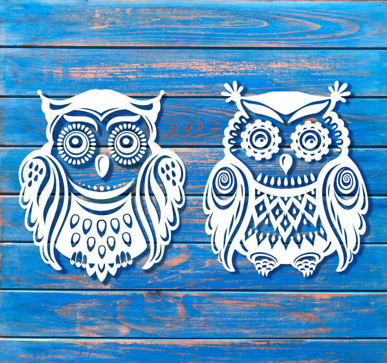 May include: Two white paper cut-out owls with intricate designs on a blue wooden background. The owls have large eyes and detailed feathers.